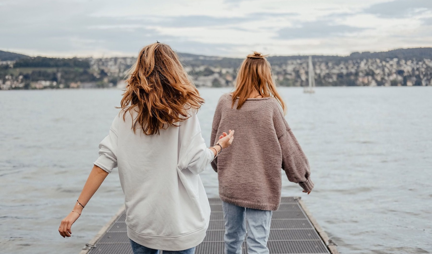 two women walk to the edge of a dock, in a body of water, looking toward the city on the other shore