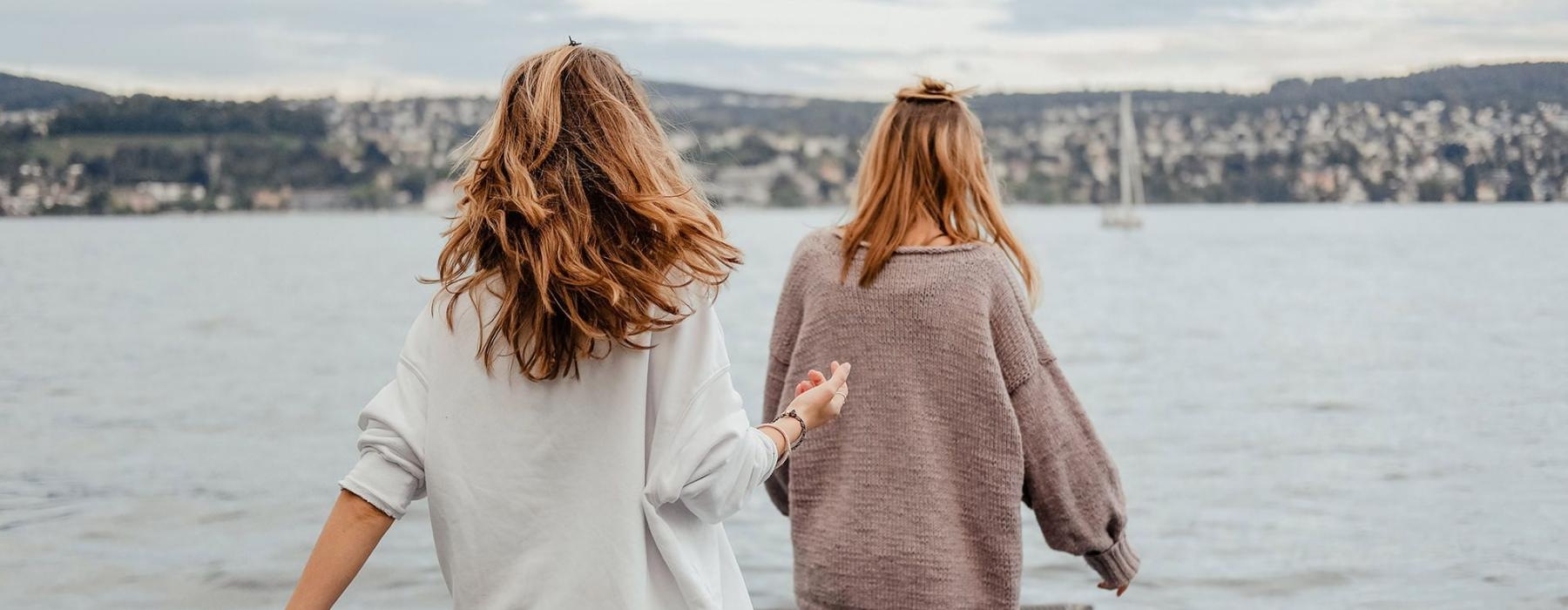 two women walk to the edge of a dock, in a body of water, looking toward the city on the other shore