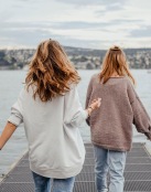 two women walk to the edge of a dock, in a body of water, looking toward the city on the other shore